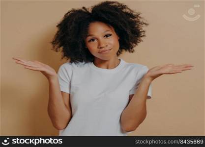 Young doubtful african woman raising palms with hesitation and looking at camera with puzzled face expression, mixed race female being unsure and having some doubts while posing against beige wall. Mixed race female being unsure and having some doubts while posing against beige wall