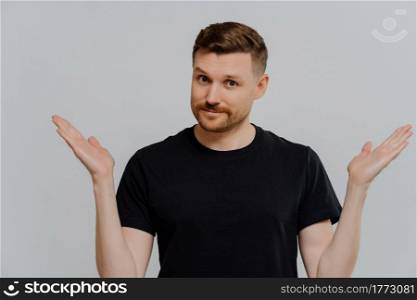 Young doubtful man in casual black t shirt cannot make decision and shrugging shoulders in uncertainty, looking at camera with facial expression of doubt and hesitation. Body language concept. Isolated shot of confused young guy spreading hands sideways and feeling doubt while making choice