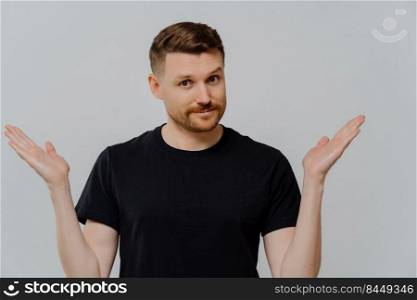 Young doubtful man in casual black t shirt cannot make decision and shrugging shoulders in uncertainty, looking at camera with facial expression of doubt and hesitation. Body language concept. Isolated shot of confused young guy spreading hands sideways and feeling doubt while making choice