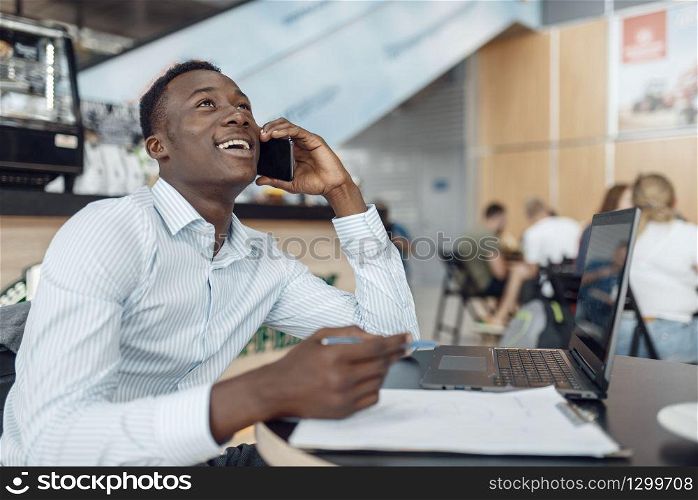 Young ebony businessman talking by phone in office cafe. Successful business person drinks coffee in food-court, black man in formal wear