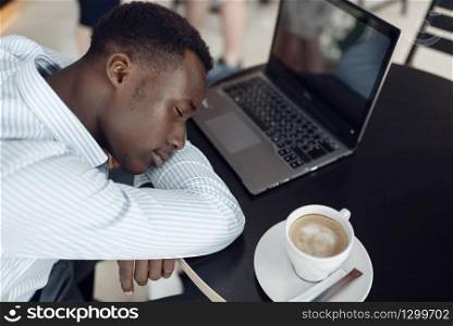 Young ebony businessman with laptop sleeping in office cafe. Tired business person drinks coffee in food-court, black man in formal wear