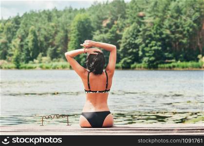 Young elegant slim tan woman in swimming bikini, sitting on the wooden jetty, lake and green forest in the background. Summer outdoor fashion, vacation, lifestyle concept. Natural light. Copy space