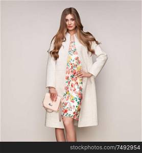 Young elegant woman in trendy white coat. Blond hair, floral dress, isolated studio shot. Fashion autumn lookbook. Model woman with handbag