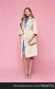 Young elegant woman in trendy white coat. Blonde hair, blue dress, isolated on pink background, studio shot. Fashion spring lookbook. Model woman with handbag