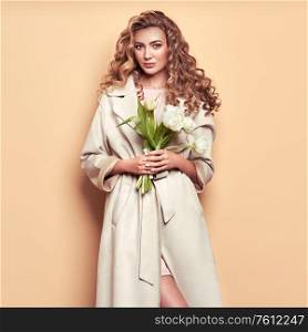 Young elegant woman in trendy white coat. Blonde hair, coral dress, isolated on beige background, studio shot. Fashion spring lookbook. Model woman with white tulips