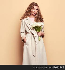 Young elegant woman in trendy white coat. Blonde hair, coral dress, isolated on beige background, studio shot. Fashion spring lookbook. Model woman with white tulips