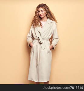 Young elegant woman in trendy white coat. Blonde hair, coral dress, isolated on beige background, studio shot. Fashion spring lookbook