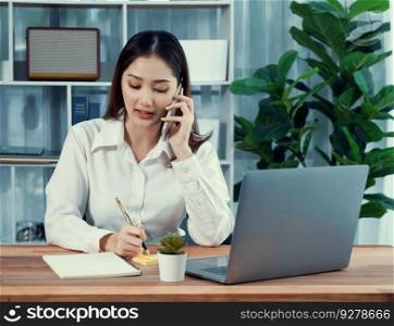 Young enthusiastic businesswoman talking on business call and writing notes on her laptop as multitask office lady. Female employee writing business tasks while talking on phone call with clients.. Young enthusiastic businesswoman talking on business call.