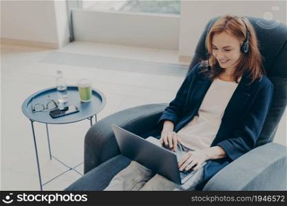 Young entrepreneur lady in headset having online conversation with partner, using laptop while sitting in armchair at home, enjoying video call on computer. Remote learning and work concept. Young entrepreneur lady wearing headset using laptop while sitting in armchair at home