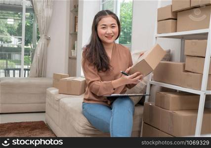 Young entrepreneur, sitting on the sofa and working at home. She smiles and holds a packing box for writing customer address inbox of deliver shipment online sales. Concept of small business owner.