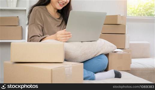 Young entrepreneur, sitting on the sofa and working at home. Checking orders from customers by laptop and writing customer address deliver shipment online sales. Concept of small business owner.