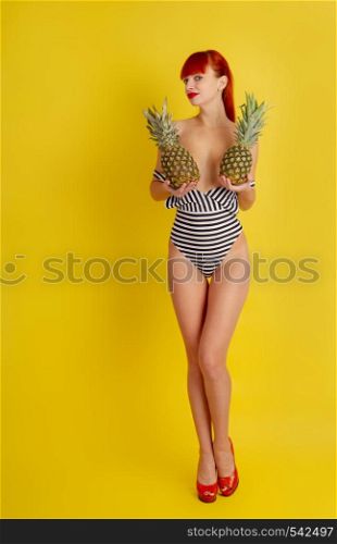 Young erotic semi-nude girl in a striped bathing suit and high-heeled shoes covers her breasts with two pineapple fruits on a bright yellow background. girl With Pineapple