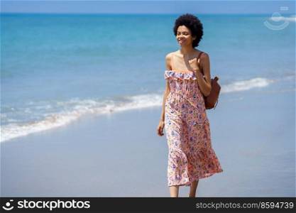 Young ethnic female with rucksack standing on beach near rippling tropical sea and looking away while travelling during summer vacation. Smiling African American woman in summer dress standing on seashore