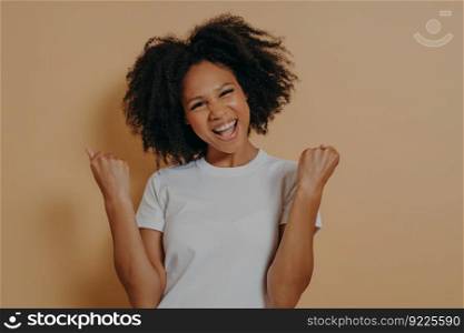 Young euphoric happy african american woman in white tshirt clenching fists and celebrating achievement, overjoyed dark skinned female making yes gesture while posing isolated on yellow background. Young euphoric woman clenching fists and celebrating achievement, isolated on yellow background