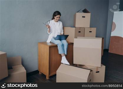 Young european business lady purchasing real estate. Businesswoman moves. Girl is sitting among cardboard containers and working on computer. Success and independence concept.. Businesswoman moves. Lady is sitting among cardboard containers and working on computer.