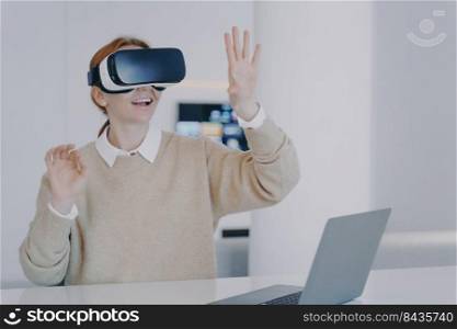 Young european girl in VR headset in office. Excited girl is sitting in front of laptop and gesturing. Designer working on project in cyberspace and concentrated on her vision. Futuristic remote work.. Young european girl in VR headset is gesturing concentrated on her vision. Futuristic remote work.