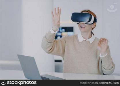 Young european girl in VR headset in office. Excited girl is sitting in front of laptop and gesturing. Designer working on project in cyberspace and concentrated on her vision. Futuristic remote work.. Young european girl in VR headset is gesturing concentrated on her vision. Futuristic remote work.