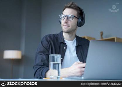 Young european man in wireless headphones listening audio podcast or watching webinar on laptop while sitting at home , german guy using computer for elearning and distant education. Young european man in wireless headphones listening audio podcast or watching webinar on laptop