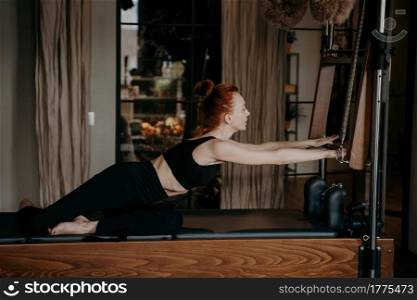 Young european redhead woman in sportswear with athletic body shape stretching arms by doing exercise on cadillac reformer in fitness studio, wearing black sportswear. People and sport. Fit young pilates instructor stretching arms on cadillac reformer