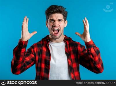 Young european stressed man in red shirt shouting isolated over blue background. Stressed and depressed guy loudly screaming to camera. Young european stressed man in red shirt shouting isolated over blue background