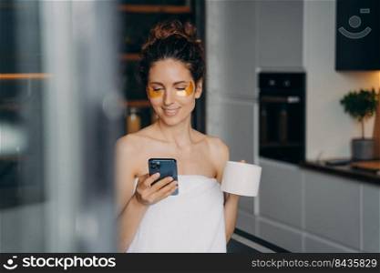 Young european woman applies eye patches and relaxing at spa hotel. Girl is messaging on phone. Happy girl wrapped in towel after bodycare beauty procedures. Modern interior of apartment.. Young european woman applies eye patches and relaxing at spa hotel. Girl is messaging on phone.