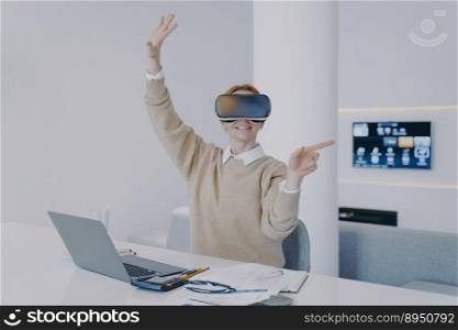 Young european woman in VR glasses clicks virtual buttons. Girl is sitting at the desk in front of laptop in office. Businesswoman working on project in cyberspace. Concept of interactive technology.. Young european woman in VR glasses click virtual buttons. Concept of technology innovation.