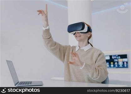 Young european woman in VR goggles in office. Student girl is sitting at the desk in front of laptop. Businesswoman working on design project in cyberspace. Futuristic remote work. Digital innovation.. Young european woman in VR goggles in office. Student is working on futuristic design in cyberspace.
