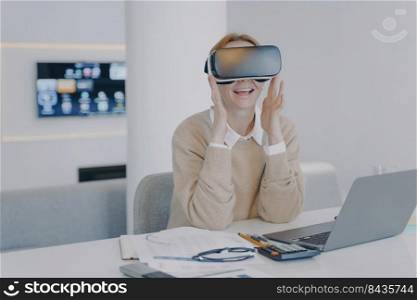 Young european woman in VR headset in office. Excited girl student is sitting at the desk in front of laptop. Concept of interactive innovation and futuristic university. Modern device using.. Excited girl in VR headset sitting at the desk in front of laptop. Working on project in cyberspace.