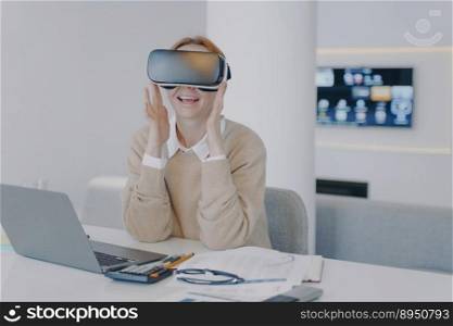 Young european woman in VR headset in office. Excited girl student is sitting at the desk in front of laptop. Concept of interactive innovation and futuristic university. Modern device using.. Excited girl in VR headset sitting at the desk in front of laptop. Working on project in cyberspace.