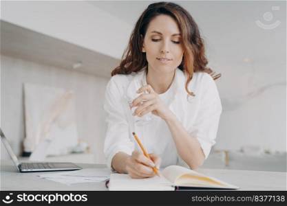 Young european woman in white is taking notes. Freelancer or businesswoman is checking schedule. Happy girl is writing down into notebook at the desk. Remote work from home on quarantine.. Young european woman in white is taking notes. Happy girl is writing down into notebook.