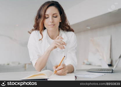 Young european woman in white is taking notes. Freelancer or businesswoman is checking schedule. Happy girl is writing down into notebook at the desk. Remote work from home on quarantine.. Young european woman in white is taking notes. Happy girl is writing down into notebook.