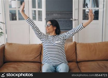 Young european woman is raising hands. Mixed race lady is stretching after daytime nap on couch. Happy woman in glasses is having rest at home enjoying weekend morning. Leather sofa in living room.. Young woman raising hands. Lady is stretching after daytime nap on couch enjoying weekend morning.