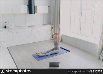 Young european woman is teaching yoga on video. Coach on exercise mat doing workout sitting on floor and looking into laptop screen. Concept of remote work and sport online on quarantine.. Young woman is teaching yoga on video with laptop. Concept of remote work on quarantine.