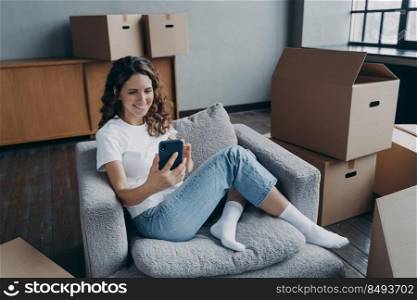 Young european woman posting news and smiling. Happy apartment buyer among boxes. Satisfied young woman is moving to new home. Girl clicks phone and chatting. Independence and achievement concept.. Happy apartment buyer among boxes. Young european woman posting news and smiling.