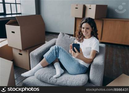 Young european woman posting news and smiling. Happy apartment buyer among boxes. Satisfied young woman is moving to new home. Girl clicks phone and chatting. Independence and achievement concept.. Happy apartment buyer among boxes. Young european woman posting news and smiling.