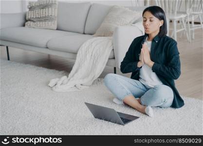 Young european woman practicing yoga online at home. Girl sitting on floor in front of laptop in lotus pose and meditating. Music listening, tranquility, stress relief. Remote lesson on quarantine.. Young woman practicing yoga online at home. Girl sitting at laptop in lotus pose and meditating.