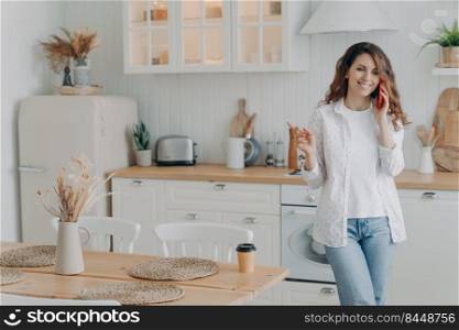 Young european woman talking on smartphone in her stylish kitchen at home. Happy attractive housewife has leisure. Modern white scandinavian interior. Stove, worktop and cuisine.. Happy housewife has leisure and talking on smartphone in kitchen. Modern scandinavian interior.