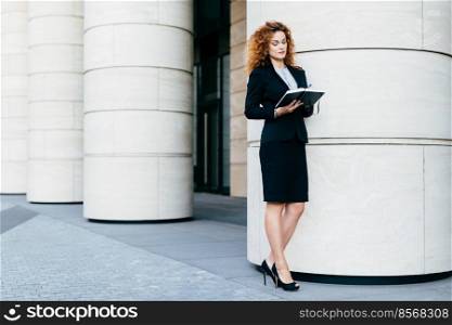 Young European woman with curly hair, wearing black formal costume and high-heeled shoes, holding her diary, writing notes, having concetrated look. Busy businesswoman making business arrangement