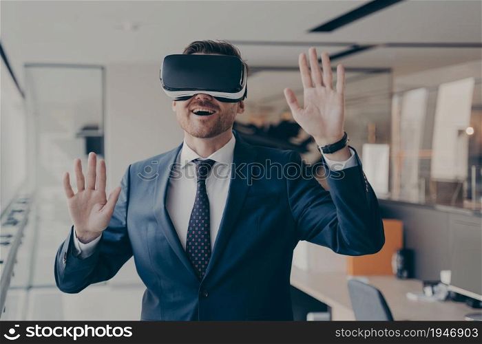 Young excited businessman dressed formally using virtual reality headset while working in office, trying vr goggles, gesturing with impressed face expression, office worker playing video games at work. Young excited businessman dressed formally using virtual reality headset while working in office