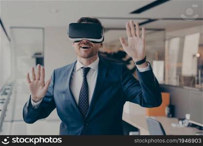 Young excited businessman dressed formally using virtual reality headset while working in office, trying vr goggles, gesturing with impressed face expression, office worker playing video games at work. Young excited businessman dressed formally using virtual reality headset while working in office