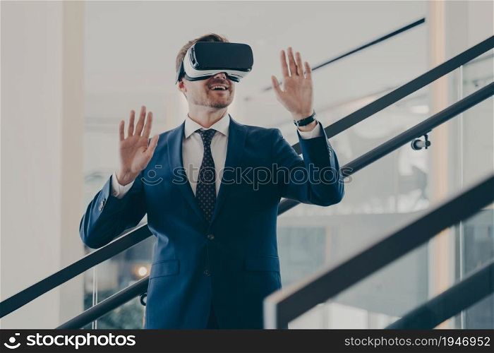 Young excited europian businessman in formal wear standing in office interior using VR headset and interacting with virtual reality simulation, testing 3d goggles at work. Cyberspace experience. Young excited europian businessman in formal wear standing in office interior using VR headset