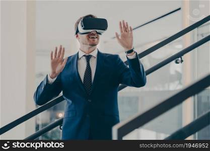 Young excited europian businessman in formal wear standing in office interior using VR headset and interacting with virtual reality simulation, testing 3d goggles at work. Cyberspace experience. Young excited europian businessman in formal wear standing in office interior using VR headset