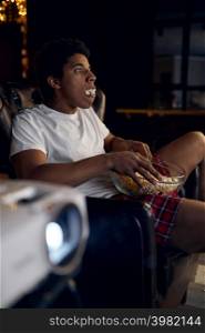 Young excited guy with big bowl of popcorn watching movie. Home cinema projector and relax time. Guy with bowl of popcorn watching movie