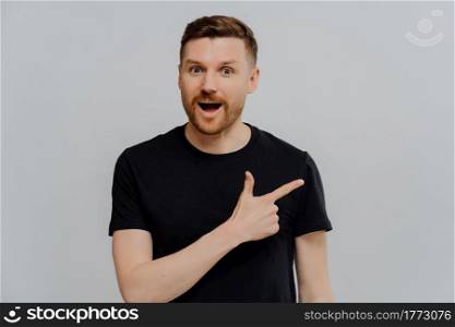 Young excited impressed man in black casual shirt pointing at upper right corner and looking at camera with surprised expression while standing isolated over grey background. Promotion concept. Amazed man pointing at upper right corner, found out special offer
