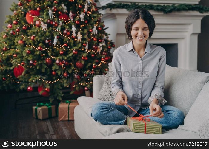 Young excited Italian woman smiling playfully with closed eyes trying to unpack Christmas gift holding ribbon with fingers while sitting on comfy sofa at home next to lush beautiful festive Xmas tree. Excited Italian woman with closed eyes trying to unpack Christmas gift while sitting on sofa at home