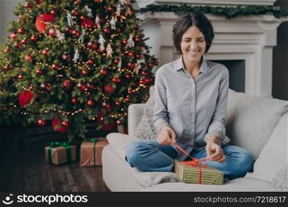 Young excited Italian woman smiling playfully with closed eyes trying to unpack Christmas gift holding ribbon with fingers while sitting on comfy sofa at home next to lush beautiful festive Xmas tree. Excited Italian woman with closed eyes trying to unpack Christmas gift while sitting on sofa at home