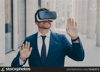 Young excited man in suit standing on city street and testing VR glasses or 3d goggles, impressed cheerful businessman using VR technology for the first time, gesturing and touching objects in air. Young excited man in suit standing on city street and testing VR glasses or 3d goggles