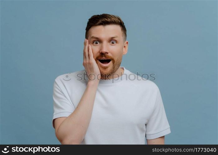 Young excited man in white shirt looking at camera with surprised face expression, keeping mouth opened while getting positive pleasant news, studio portrait of amazed man. Human emotions concept. Young excited man hearing positive pleasant news and being in shock