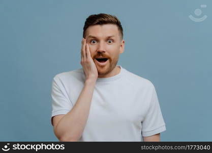 Young excited man in white shirt looking at camera with surprised face expression, keeping mouth opened while getting positive pleasant news, studio portrait of amazed man. Human emotions concept. Young excited man hearing positive pleasant news and being in shock