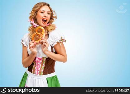 Young excited Oktoberfest woman wearing a traditional Bavarian dress dirndl holding a pretzel in hands on blue background.